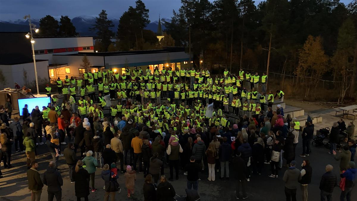 Mange barn med gule refleksvester står oppstilt i et utendørs amfi foran mange foresatte. Foto - Klikk for stort bilde